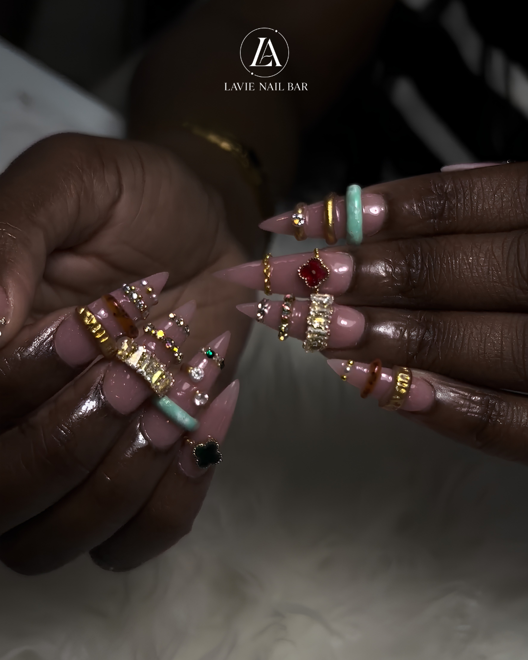 Long stiletto nude nails adorned with gold bands, crystal rows, and red and green clover charms
