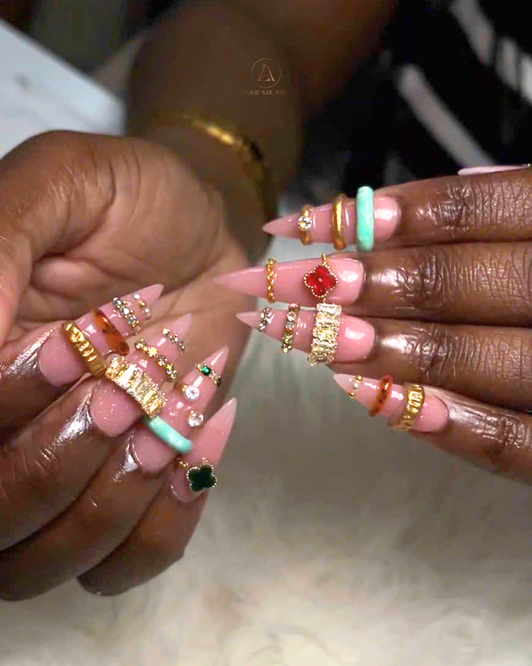 Long stiletto nude nails adorned with gold bands, crystal rows, and red and green clover charms